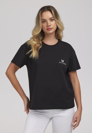 Młoda kobieta z długimi falującymi blond włosami, ubrana w czarną koszulkę Lee Cooper i białe spodnie, stoi z jedną ręką na biodrze na jednolitym tle.