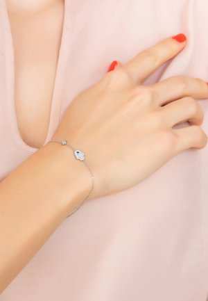 Silver bracelet featuring a detailed hamsa charm with blue accents and a small round bead, worn on a hand resting against a soft pink surface.
