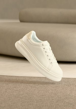 Baskets basses blanches avec lacets sur un tapis beige, placées devant un canapé marron clair.