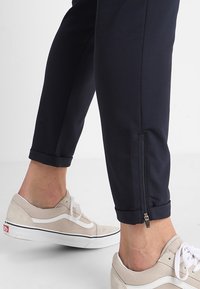 Marina byxor med ett strukturerat mönster och blixtlås vid anklarna, i kombination med ljusgrå skor med vita detaljer och en svart rand.