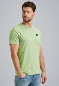 Camiseta verde claro de manga corta en material de algodón suave, con un pequeño parche de logo negro en el área del pecho izquierdo.