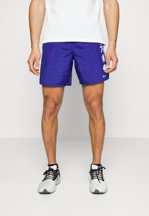 Homme portant un short de sport violet avec des motifs abstraits blancs, un t-shirt de sport blanc, et des baskets blanches avec des lacets noirs sur un fond uni.