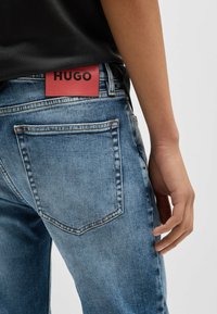 Jeans in denim leggero blu chiaro con una finitura sbiadita. Presentano una toppa in pelle rossa con l'etichetta "HUGO" in vita e due tasche posteriori.