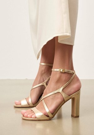 Pieds portant des sandales à talons hauts dorées à brides avec boucle à la cheville, associées à un pantalon large longueur cheville de couleur blanc cassé.