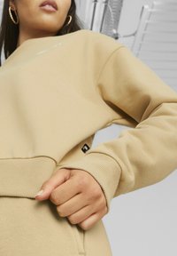 Felpa beige con polsini e orlo a coste, texture morbida, con un'etichetta del logo visibile vicino alla tasca. Primo piano della mano nella tasca.