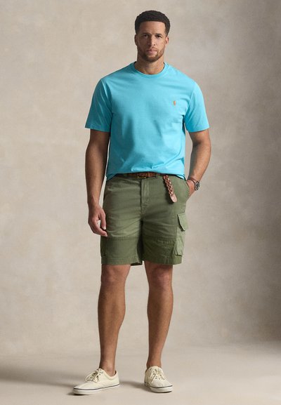 Ανοιχτό μπλε βαμβακερό μπλουζάκι, πράσινα cargo σορτς με τσέπες, καφέ πλεκτή ζώνη και ανοιχτά παπούτσια· το μοντέλο στέκεται μπροστά σε ουδέτερο φόντο.