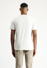 Camiseta blanca de algodón con mangas cortas, cuello redondo y una textura suave, combinada con pantalones marrones de textura. Capturada desde la parte de atrás.