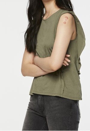 Kobieta z długimi ciemnymi włosami, ubrana w oliwkowo-zielony, bezrękawnik i ciemne jeansy, pokazująca okrągły siniak na górnej części prawego ramienia.