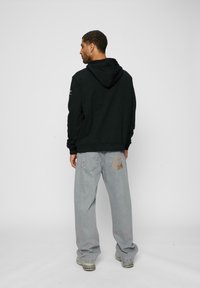Sort hættetrøje med ribbet kant. Lysegrå baggy jeans med et logo på baglommen og kontraststing. Sneakers synlige.