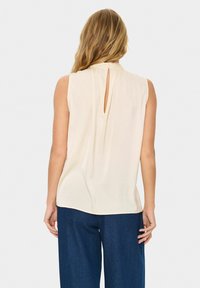 Top beige senza maniche con colletto alto e dettaglio a chiave sul retro, caratterizzato da una vestibilità comoda e una superficie liscia. Abbinato a pantaloni blu scuro.