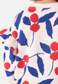 Baumwollpullover mit weißem Hintergrund und rot-blauem Blumenmuster. Rüsche am Schulterbereich. Etikett auf dem Stoff sichtbar.