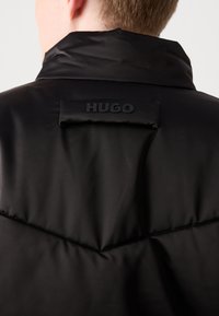 Black padded vest with high collar, featuring a textured “HUGO” logo patch on the back and a diagonal seam detail. Smooth, sleek material.