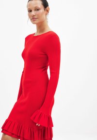 Rode geribbelde gebreide jurk met lange mouwen en uitlopende mouwen, met een ruches aan de onderkant. Glad textuur, aansluitend ontwerp.