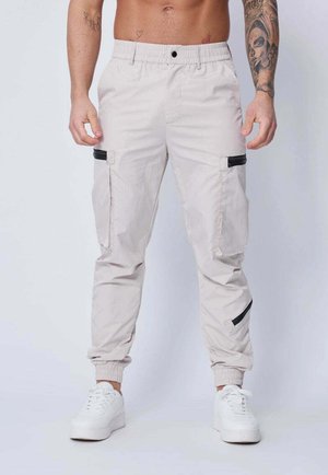 Pantalons cargo beiges avec coupe fuselée, taille élastique et chevilles resserrées. Dotés de poches zippées noires pour le rangement. Tissu lisse et léger.