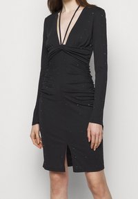 Robe noire à manches longues avec un décolleté en V profond, des détails froncés et une fente sur le devant. Embellie de petits accents métalliques sur toute la surface.