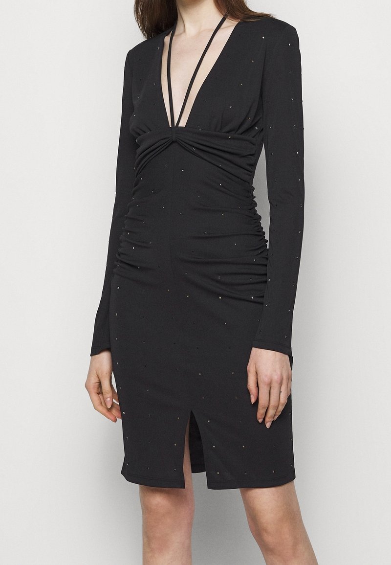 Robe noire à manches longues avec un décolleté en V profond, des détails froncés et une fente sur le devant. Embellie de petits accents métalliques sur toute la surface.