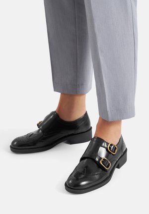 Zapatos de cuero negro con acabado brillante, diseño de puntera alada, dos hebillas decorativas, tacón bajo de bloque, combinados con pantalones sastre gris claro.