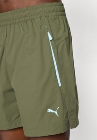 Olivinės sportinės šortai su elastine juosta, šviesiai mėlyna užtrauktuku užsegama kišenė ir Puma logotipas ant kairiojo šlaunies, dėvimi žmogaus.