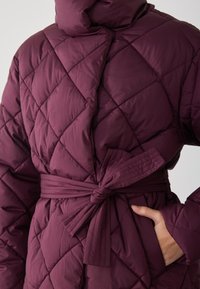 Cappotto imbottito bordeaux trapuntato con cappuccio, vita regolabile, tessuto texturizzato e motivo a cuciture diagonali con dettagli delle tasche laterali.