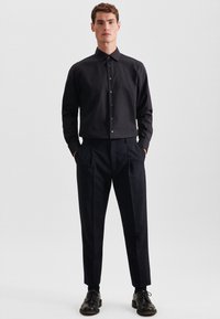 Uomo con camicia nera abbottonata, pantaloni su misura neri e scarpe eleganti nere, in piedi con le mani in tasca davanti a uno sfondo semplice.