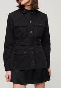 Chaqueta de lienzo negra con cierre de botones, cuatro bolsillos frontales y cintura ceñida con cinturón. Presenta un cuello y mangas largas. Tejido texturizado.