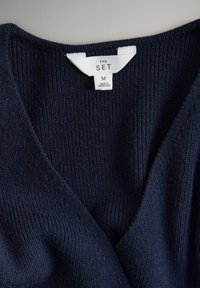 Vêtement tricoté côtelé bleu foncé avec une étiquette blanche portant la mention « THE SET M Made in Bangladesh » près du col.