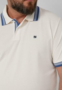 Jasno-beżowa koszulka polo o teksturowanym wykończeniu, z niebieskimi paskami na kołnierzyku i małym logo na piersi. Posiada dwa guziki przy szyi.