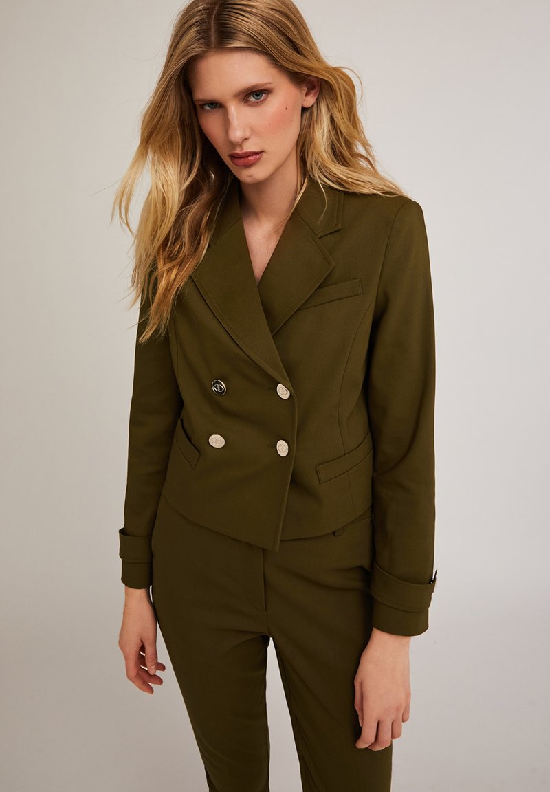 Blazer doppiopetto verde oliva con bottoni argentati, taglio su misura, revers strutturati e tasche laterali. Tessuto liscio con leggera lucentezza.
