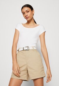 White short-sleeve fitted top paired with beige cotton shorts and a white belt featuring a decorative silver buckle. Smooth textures throughout.