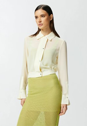 Blusa ligera amarilla pálido con frente abotonado y cuello; combinada con una falda de tejido verde lima con un patrón de zigzag, que muestra un corte ajustado.