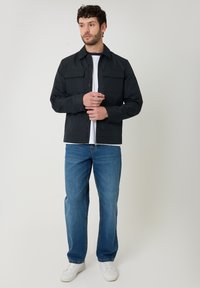 Hombre con chaqueta negra, camisa blanca, vaqueros azules y zapatillas blancas, de pie ajustando la cremallera de la chaqueta sobre un fondo liso.