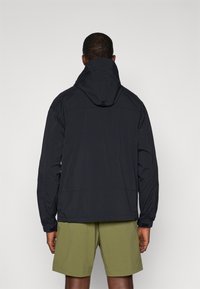 Veste imperméable noire avec un col haut et une capuche ajustable, dotée de manches longues et de poignets élastiques, portée par-dessus un short vert olive.