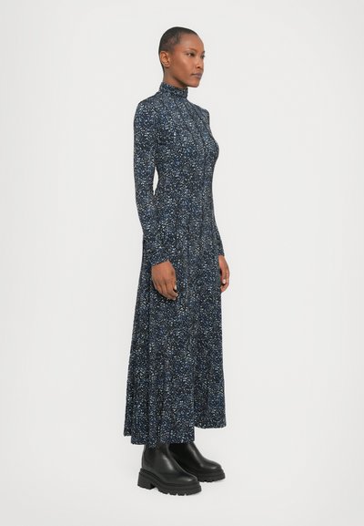 Long-sleeve, high-neck dress in navy with a speckled pattern. Flowing silhouette with a fitted bodice and textured fabric, paired with black boots.