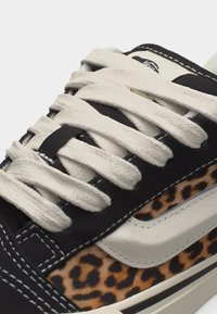 Κοντινό πλάνο αθλητικού παπουτσιού με κορδόνια σε κρεμ χρώμα, μαύρες και λευκές επιφάνειες, και λεπτομέρεια με σχέδιο λεοπάρ κατά μήκος της πλευράς.