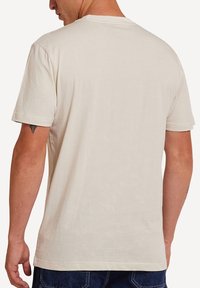 Cream cotton t-shirt with short sleeves, a crew neck, and a plain, smooth texture. Includes a visible tattoo on the left arm.