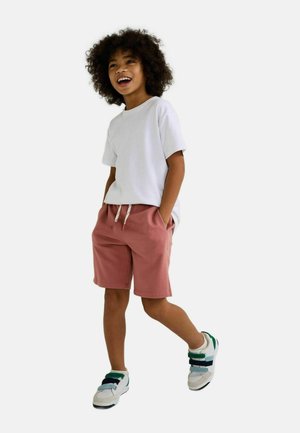 Enfant souriant marchant les mains dans les poches, portant un t-shirt blanc, un short rose et des baskets blanches à lanières vertes et bleues.