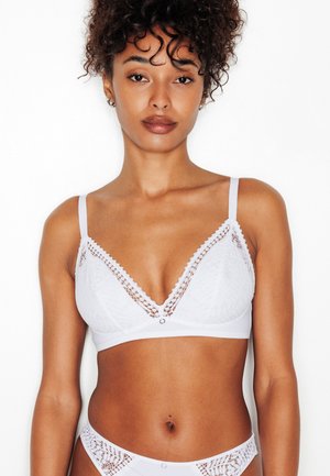 Femme aux cheveux bouclés portant un bralette en dentelle blanche et une culotte assortie, debout devant un fond blanc uni.