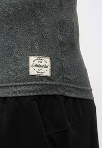 Γκρι υφασμάτινο μπλουζάκι Nike με λευκή ραμμένη ετικέτα που γράφει "Established 1972 Nike Athletic Club Just Do It" πάνω από μαύρο παντελόνι.