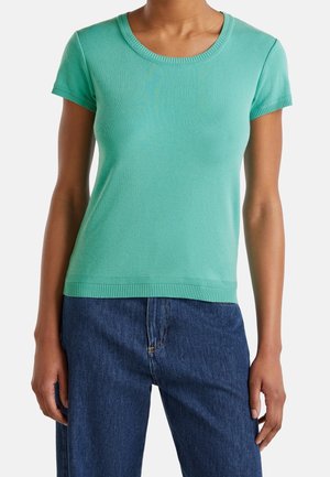 Person wearing a fitted short-sleeve turquoise sweater and high-waisted dark blue jeans standing against a plain background.