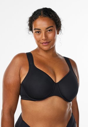 Femme aux cheveux bouclés portant un soutien-gorge noir avec armatures, souriante et face à un fond blanc uni.