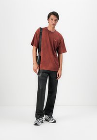 En ung mann iført en slitt rød oversized t-skjorte, svarte skinnbukser, sølvfargede sneakers og en svart crossbody-veske, står mot en hvit vegg.