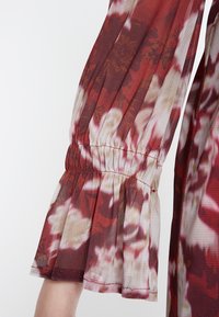 Ärmel eines Blumenkleides aus leichtem Stoff in Rot-, Weiß- und Beigetönen; mit gerafftem, elastischem Bund und Rüschendetail.