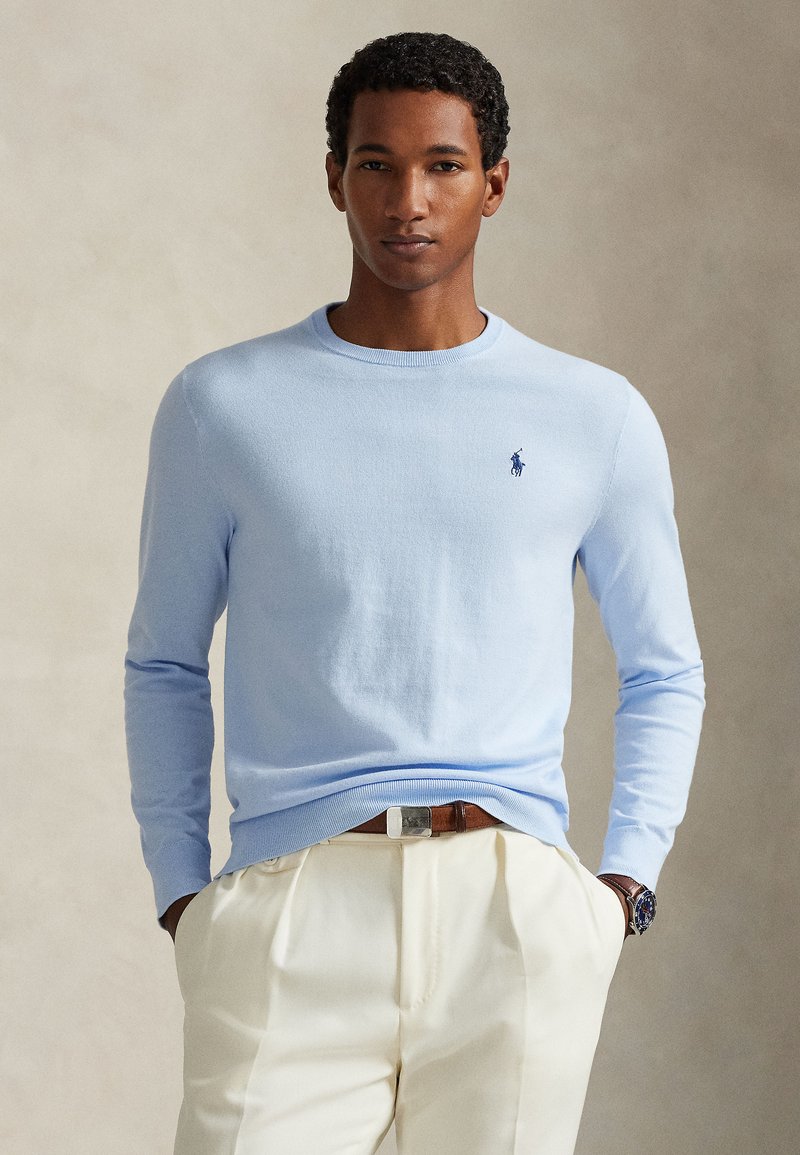 Maglione azzurro chiaro a maniche lunghe in maglia con scollo rotondo e piccolo logo scuro. Indossato con pantaloni color crema e cintura marrone.