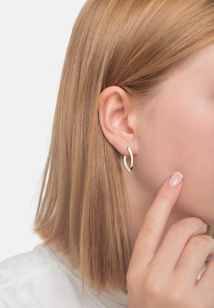 Złote kolczyki w kształcie obręczy o teksturowanej powierzchni, o okrągłym kształcie i otwartym wzorze, noszone w lewym uchu; krótkie, proste włosy okalające twarz.