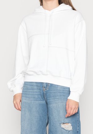 Kvinde iført en hvid hættetrøje med snøre og lyseblå revnede jeans, stående mod en ensfarvet baggrund.