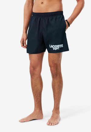 Hombre que lleva puestos unos pantalones cortos de baño negros Lacoste, de pie descalzo con una mano en el bolsillo sobre fondo blanco.