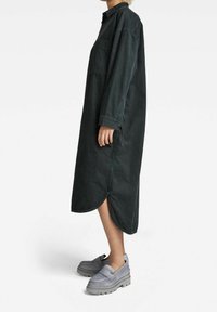 Robe chemise longue vert foncé en denim, avec col, poches latérales et ourlet arrondi. Associée à des chaussures épaisses grises.