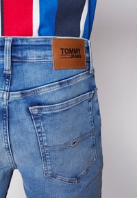 Vista trasera de unos jeans de mezclilla de color azul claro con un parche de cuero marrón de Tommy Jeans y una pequeña bandera de Tommy Hilfiger en el bolsillo trasero.