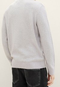Maglione grigio chiaro con texture e motivo a griglia, polsini e orlo a coste. Indossato sopra jeans neri. La vista da dietro mostra la vestibilità e il design complessivo.