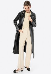 Cappotto lungo in pelle nera, top beige, pantaloni su misura chiari e scarpe con tacco a blocco nere e beige con dettaglio a fiocco.